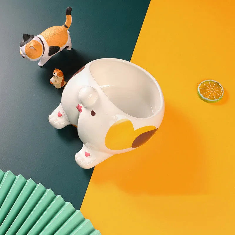 Ceramic pet bowl with inclined mouth and cartoon cat butt design, in white, on a wooden floor.
