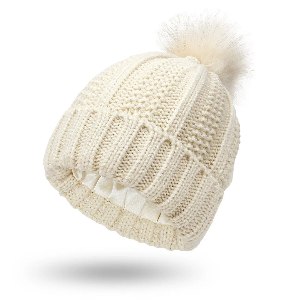 Women's black knit beanie hat with a white faux fur pom pom and satin lining for hair protection.