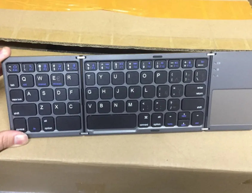 Black tri-fold folding Bluetooth keyboard with touchpad, shown both folded and unfolded next to a smartphone.