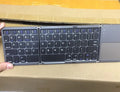 Black tri-fold folding Bluetooth keyboard with touchpad, shown both folded and unfolded next to a smartphone.