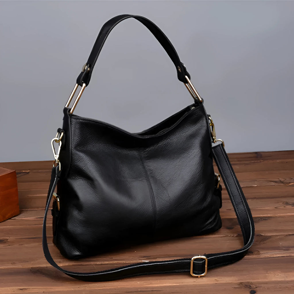A vertical square-shaped genuine leather crossbody bag in black, showing external sewing lines and a crossbody strap.