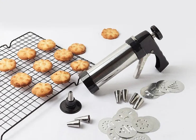 Stainless steel biscuit press gun with assorted cookie moulds and icing nozzles on a baking sheet.