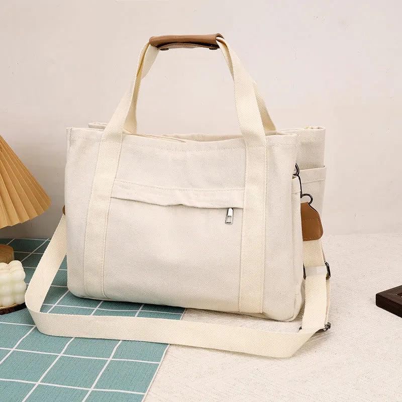 Horizontal square canvas tote bag in neutral colour with open zipper sides showing internal phone, document, computer and camera pockets